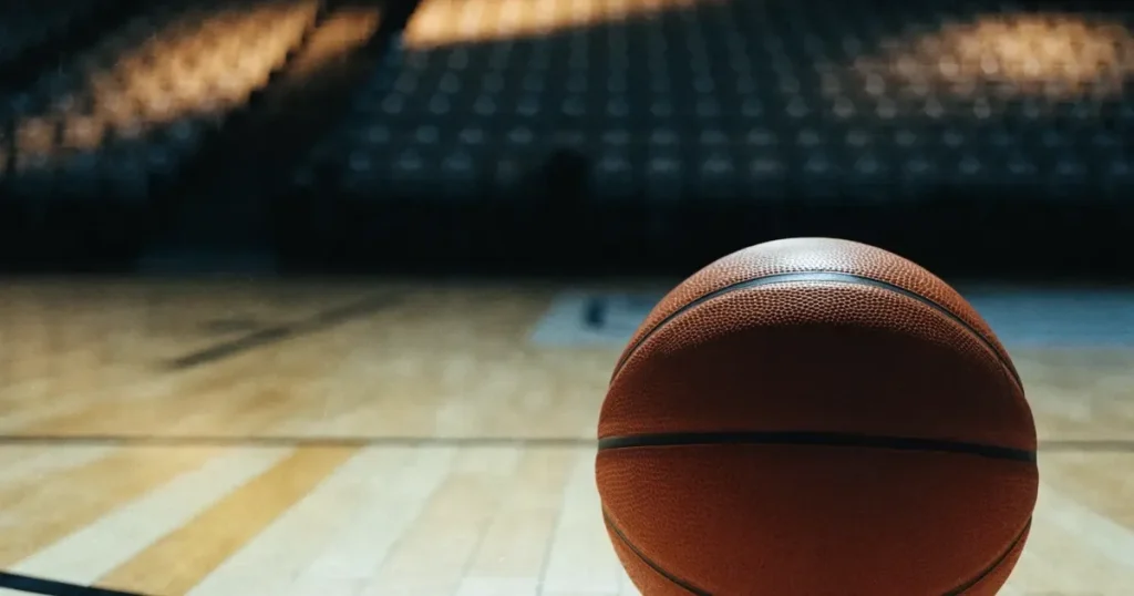 Ein Basketball liegt auf dem hellen Parkett einer leeren Halle, im Hintergrund steigen die Tribünenreihen im Halbdunkel an.