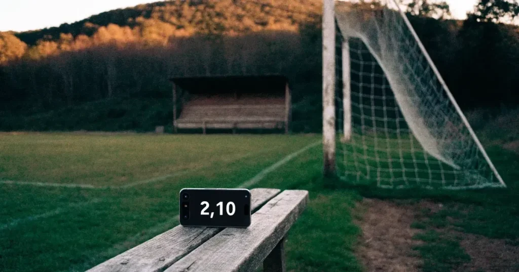 Ein kleiner Fussballplatz mit einfacher Holztribüne und Rasenstreifen am Rand einer Ortschaft, im Hintergrund ein bewaldeter Hügel im Abendlicht.