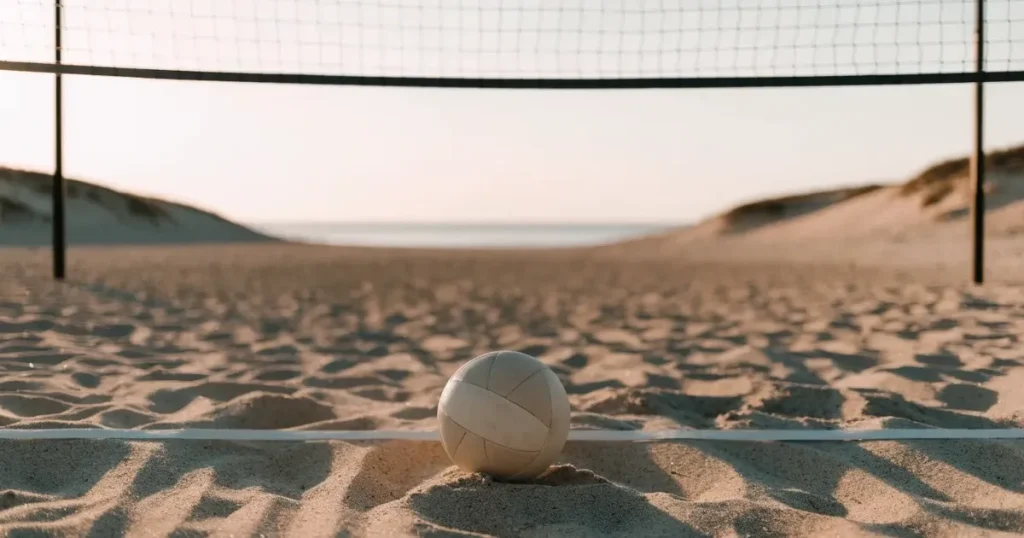 Ein Volleyball liegt im Sand eines Beachplatzes, zwischen zwei Pfosten ist ein Netz gespannt, tiefe Abendsonne wirft lange Schatten.