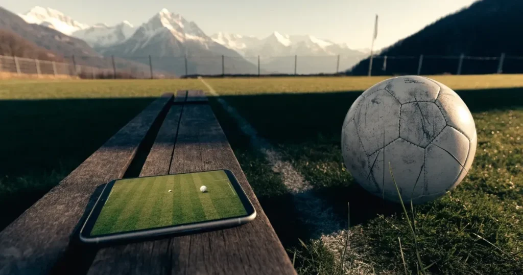 Ein Dorf-Sportplatz mit einem Ball am Strafraumrand, dahinter schneebedeckte Alpengipfel im klaren Morgenlicht